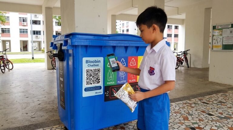 Primary school student in Singapore school uniform standing at a blue Bloobin recycling bin near an HDB void deck, holding a recyclable item and looking at the bin's information sticker